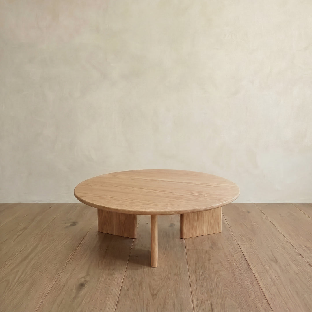 The Round coffee table in white oak features a minimalist, modern design with four sturdy legs and sits on a matching wooden floor against a plain, light-colored wall.