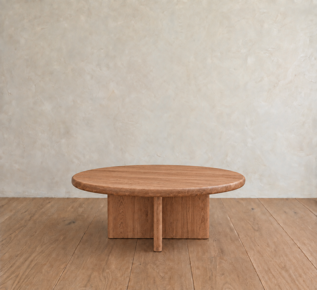 The Round coffee table in red oak features a minimalist, handmade design and rests on a wooden floor against a textured light beige wall.