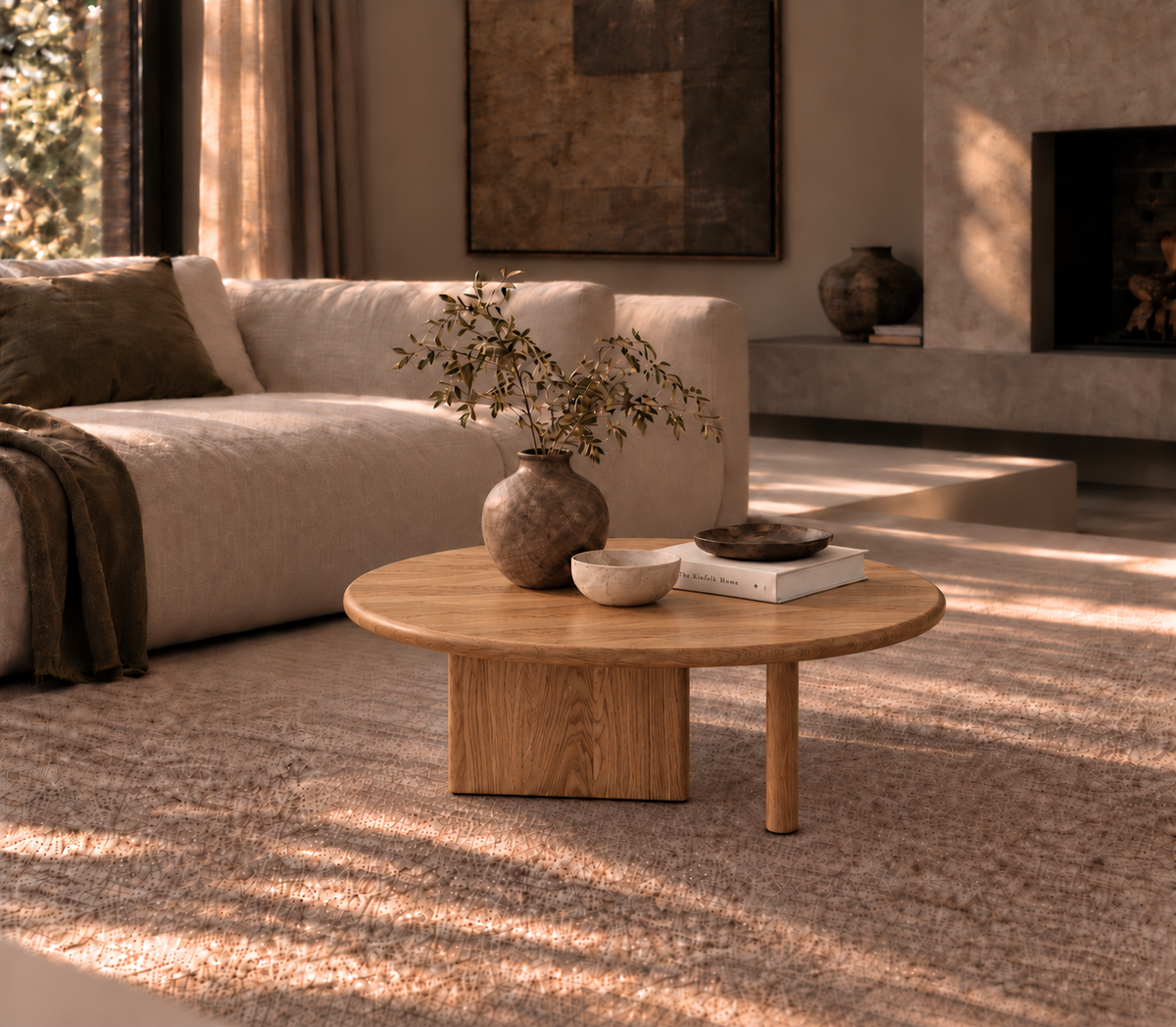 A Round coffee table in red oak, handcrafted in Vancouver, is adorned with a vase of green branches, a bowl, and two books. It rests on a beige rug in a cozy, sunlit living room featuring a modern sofa and textured decor.