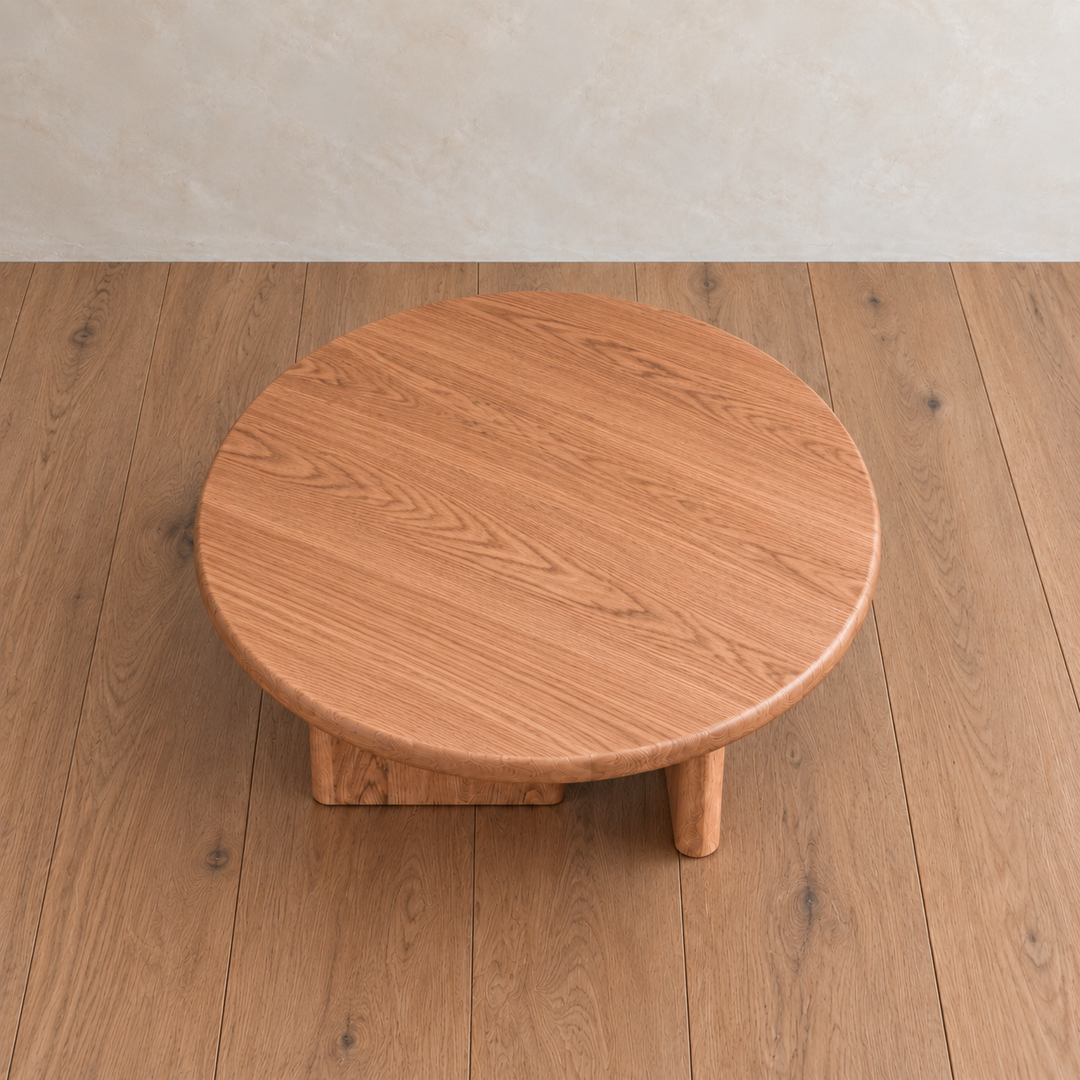 A Round coffee table in red oak, handmade with three thick legs, sits on a matching wooden floor against a light beige wall.