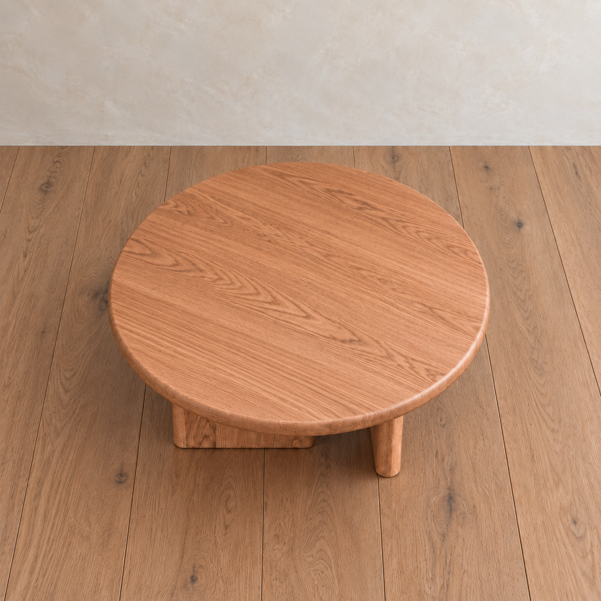 A Round coffee table in red oak, handmade with three thick legs, sits on a matching wooden floor against a light beige wall.