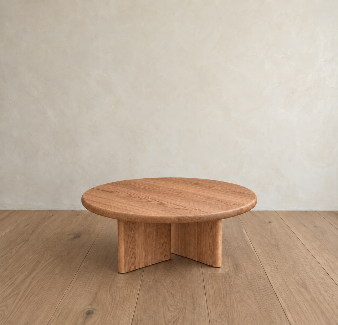 The Round coffee table in red oak, handmade in Vancouver with a minimalist design, sits on a wooden floor against a light beige textured wall.