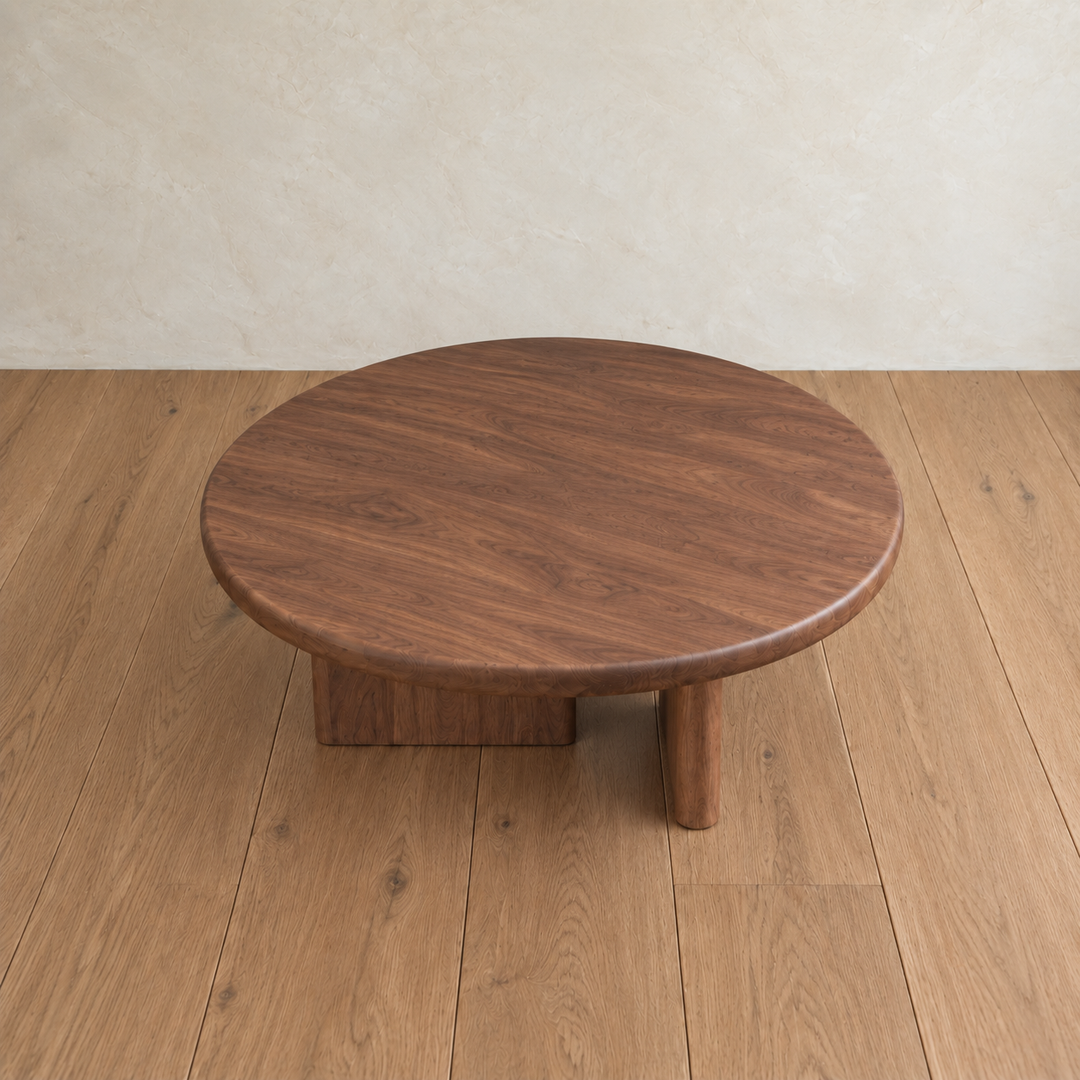 The Round coffee table in walnut, featuring a thick top and sturdy, asymmetrical legs, sits on a light wood floor against a neutral textured wall.