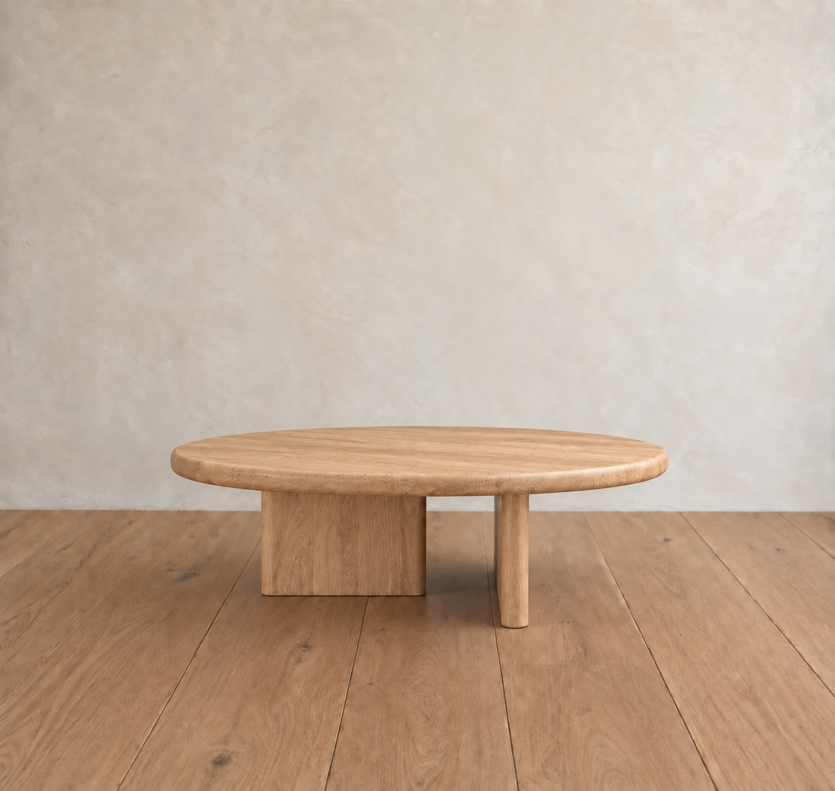 Round coffee table in white oak, handmade in Vancouver, features a unique base with one rectangular and one cylindrical leg. It sits gracefully on a wooden floor against a light textured wall.