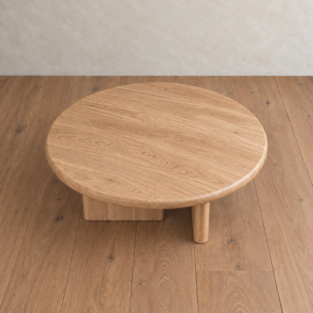 A Round coffee table in white oak, handmade in Vancouver with three legs, sits on a matching wooden floor against a beige textured wall.