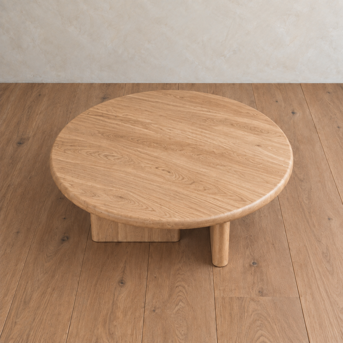 A Round coffee table in white oak, handmade in Vancouver with three legs, sits on a matching wooden floor against a beige textured wall.