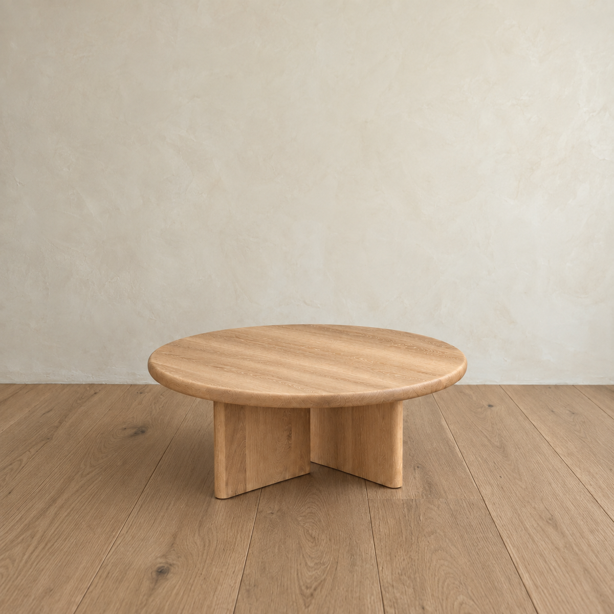 The Round coffee table in white oak, handmade in Vancouver, displays a smooth surface and intersecting panel legs as it rests on a wooden floor against a light, textured wall.