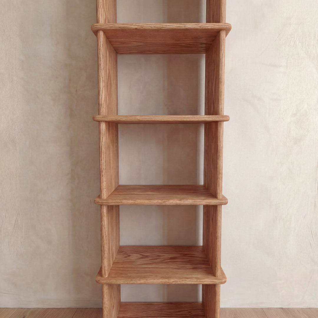 The Narrow bookshelf in red oak is a tall, handmade wooden piece with five evenly spaced shelves and a natural finish that highlights the grain. It stands empty against a light-colored textured wall.