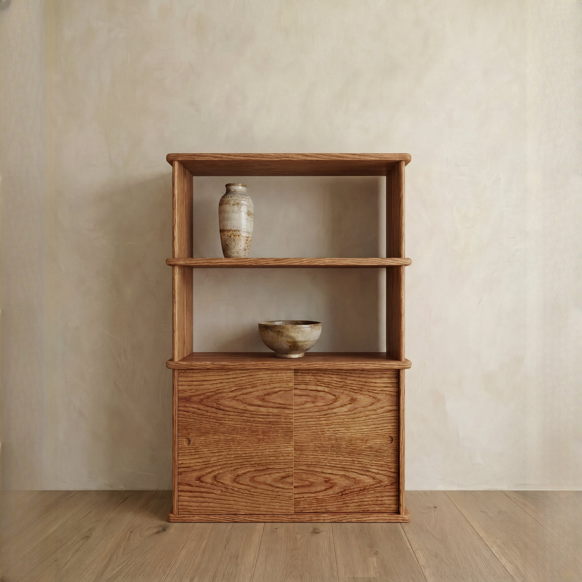 The Sliding door bookcase in red oak stands against a neutral wall and wooden floor, with two open shelves—one displaying a ceramic vase and the other a ceramic bowl.