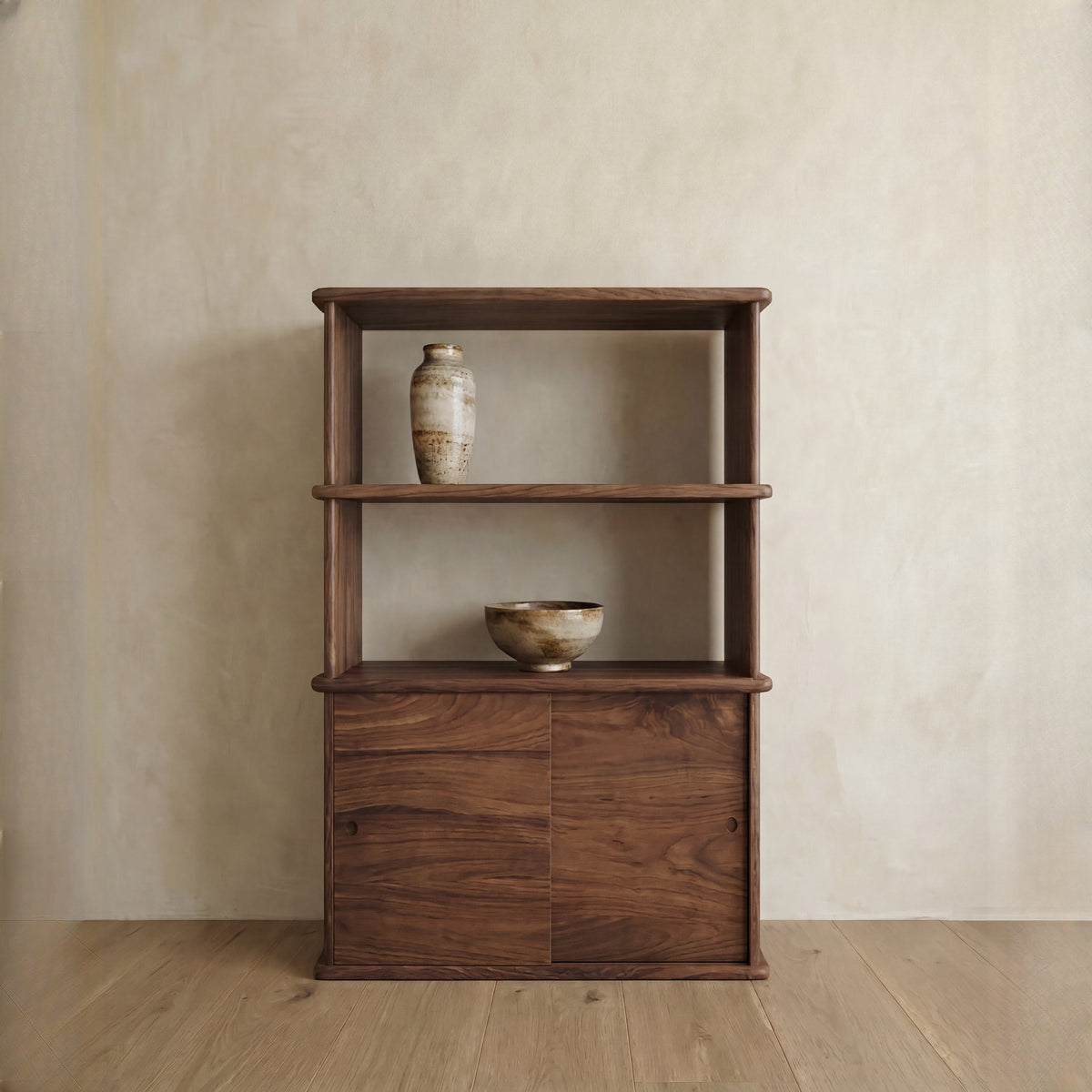 A sliding door bookcase in walnut with two shelves displays a ceramic vase on the top and a ceramic bowl on the middle shelf. Handmade in Vancouver, it stands against a light textured wall on a wooden floor.