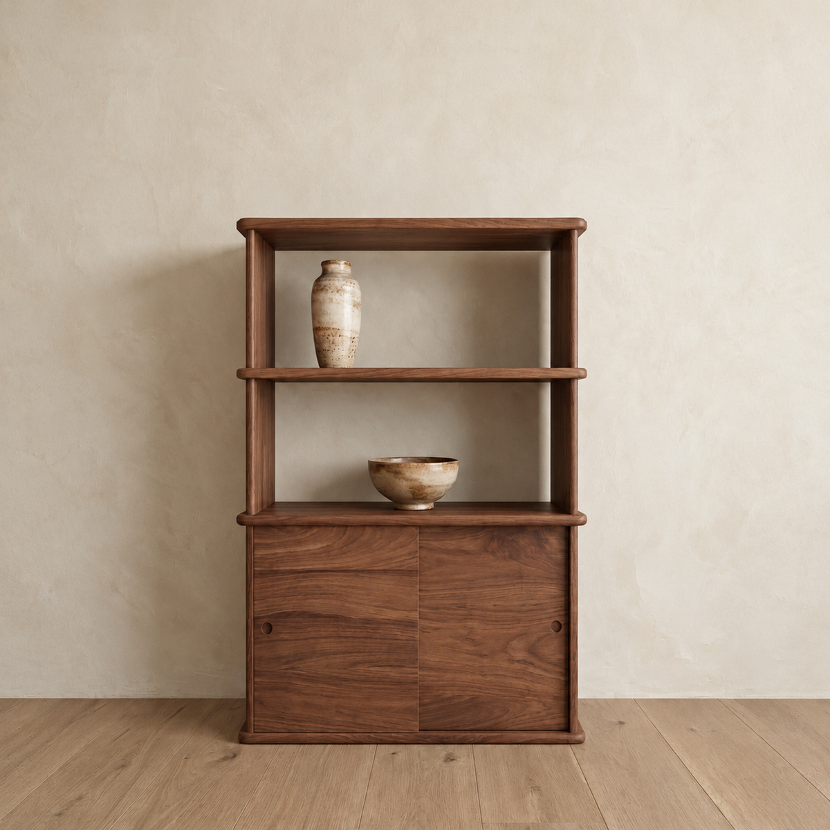 The sliding door bookcase in walnut against a plaster wall, with two open shelves above a sliding-door cabinet, holding a ceramic vase and a bowl.