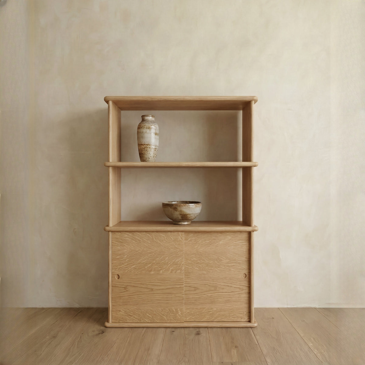 The Sliding door bookcase in white oak, handmade in Vancouver, has two shelves and a cabinet with sliding doors. A ceramic vase is on the top shelf and a ceramic bowl on the middle shelf, set against a light beige wall and wood floor.