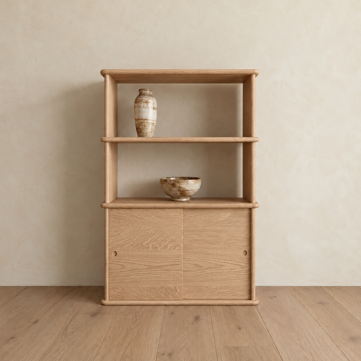 The sliding door bookcase in white oak against a plaster wall, with two open shelves above a sliding-door cabinet, holding a ceramic vase and a bowl.