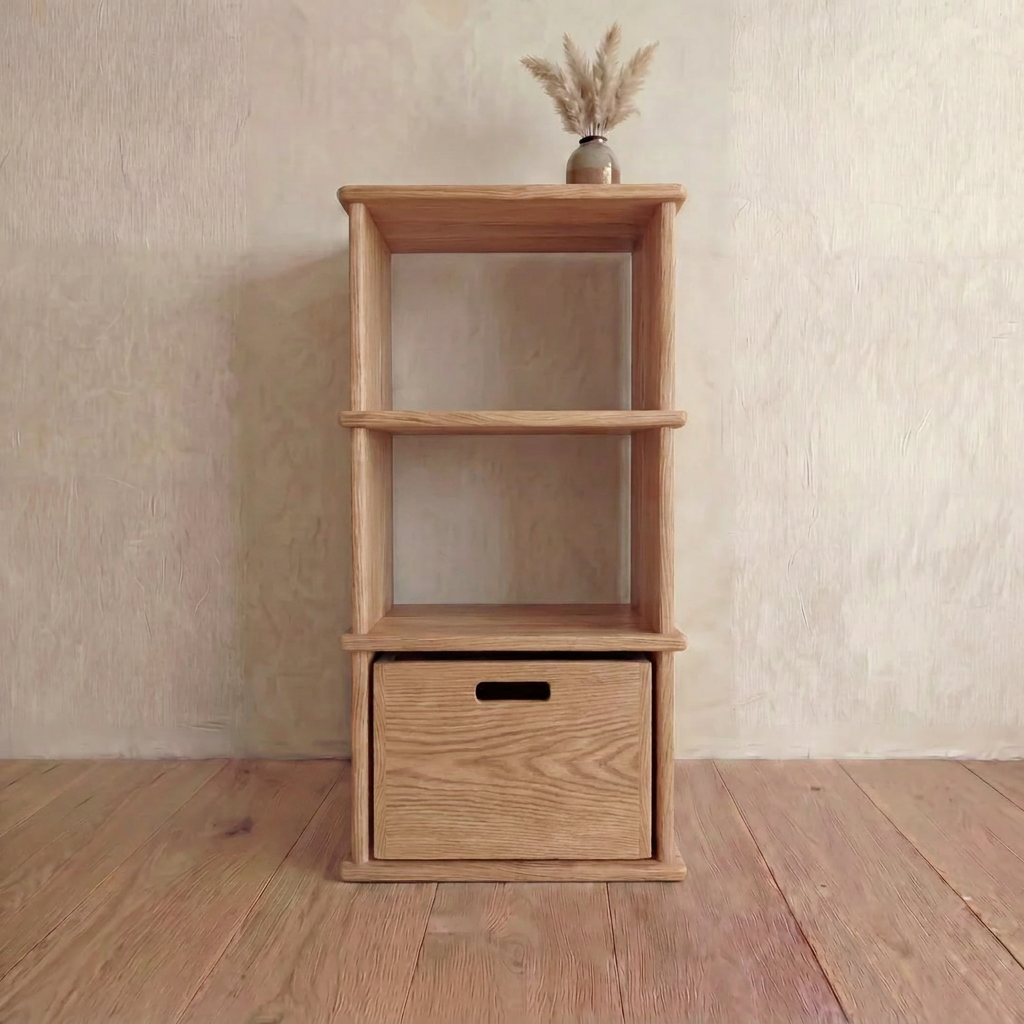 The Small bookshelf in white oak features two open shelves and a bottom drawer, crafted by hand. It stands on a wooden floor with a small vase of dried grass displayed on the top shelf.