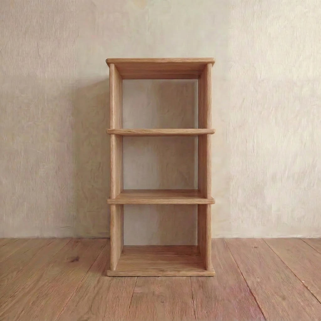 The Small bookshelf in white oak features three handmade shelves and stands against a beige wall on a wooden floor, shown in natural lighting.