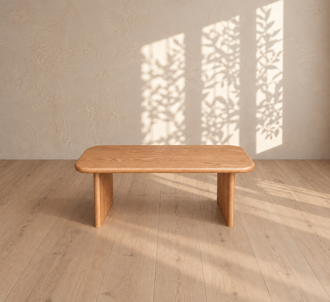 A 40 inch coffee table in red oak, handmade from solid wood, sits on a light wood floor. Sunlight streams through three windows, casting leafy shadows on a beige wall behind the table.