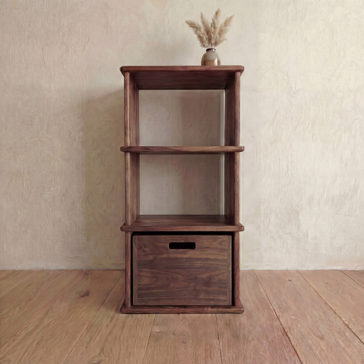 A small bookshelf in walnut with a wooden storage cube on the bottom shelf and a vase of pampas grass on top.