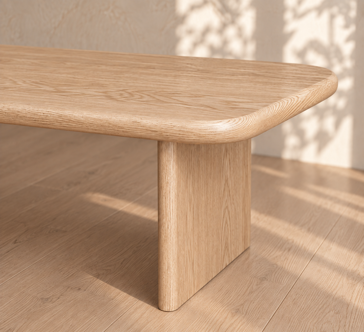 A close-up of a 40 inch coffee table in white oak, featuring a handmade, minimalist design with rounded edges and a thick, flat leg, sitting on a matching wood floor. Sunlight and leafy shadows fall across the scene.