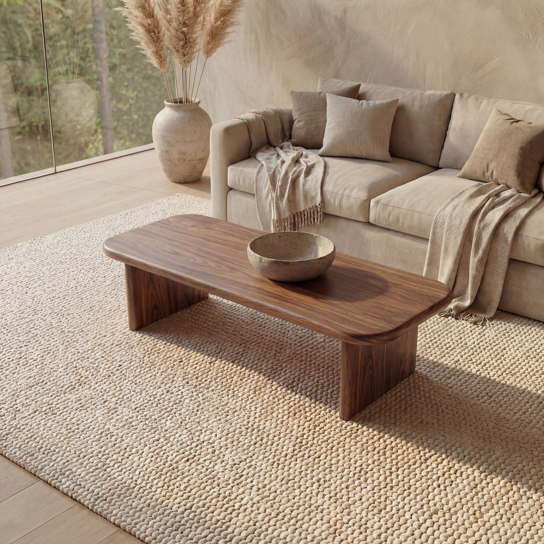 A cozy living room features a beige sofa, neutral pillows, and a throw blanket. A 60 inch coffee table in walnut with a bowl rests on a textured rug, while a large vase with pampas grass and tall windows create a serene atmosphere.