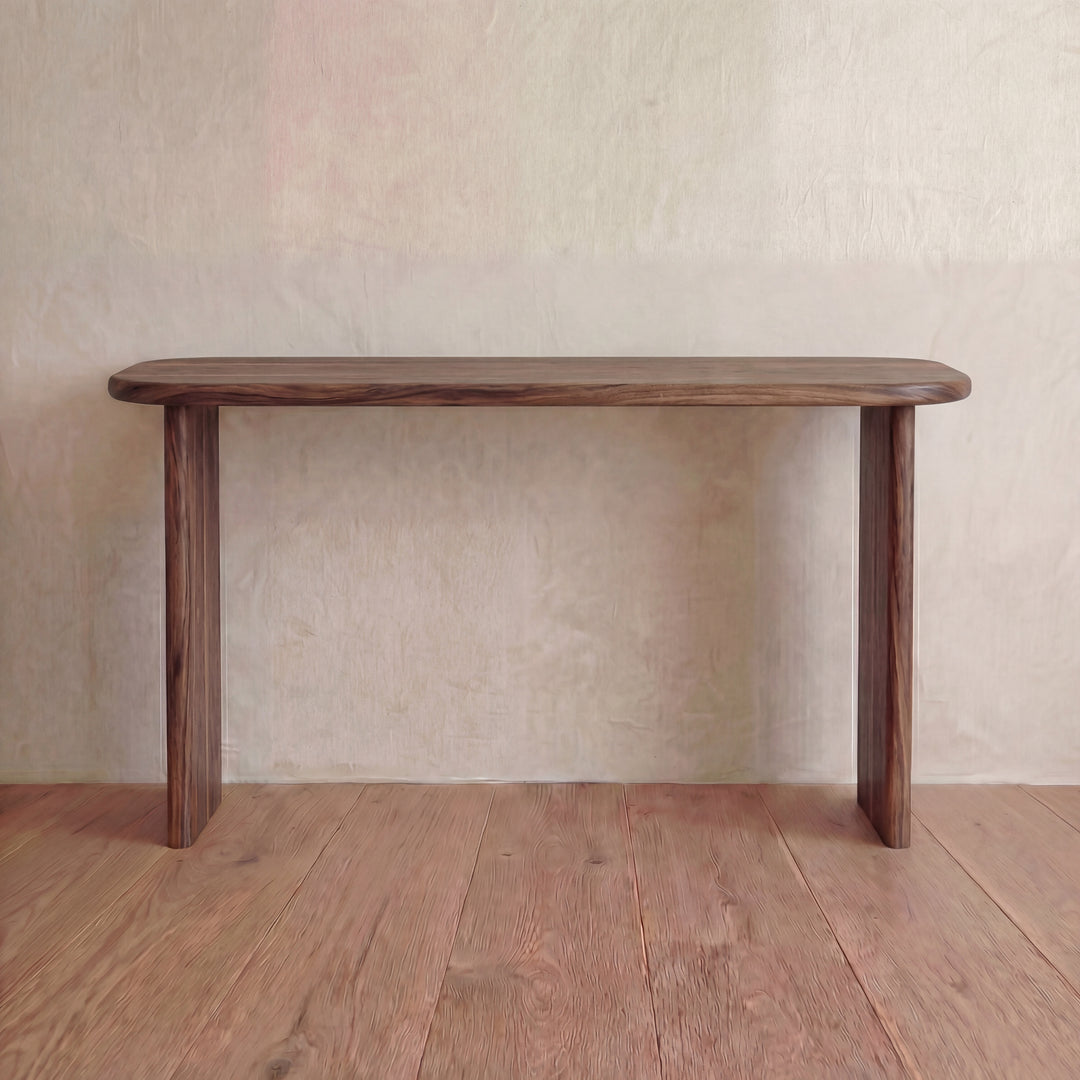 The 60 inch console table in walnut, handmade in Vancouver with rounded edges and three legs, stands against a light-textured wall on a wooden floor.