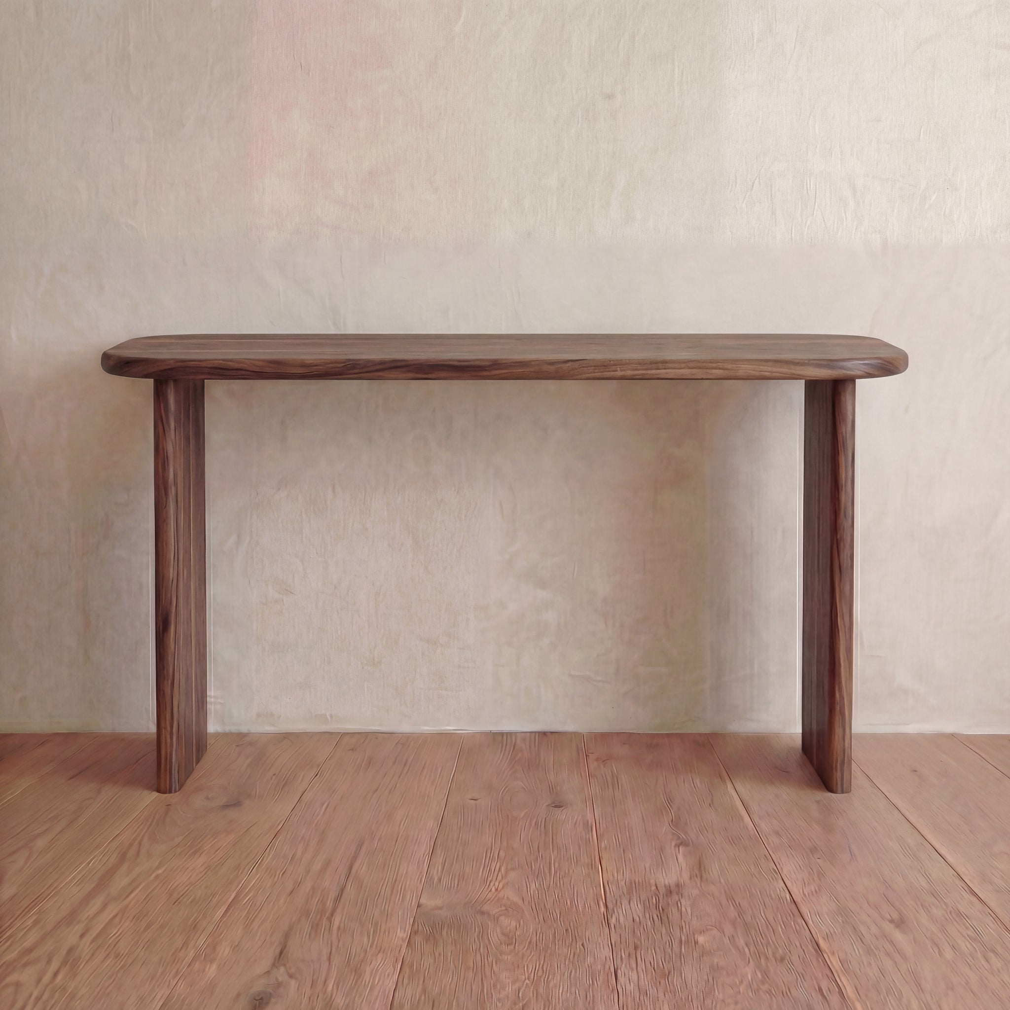 The 60 inch console table in walnut, handmade in Vancouver with rounded edges and three legs, stands against a light-textured wall on a wooden floor.