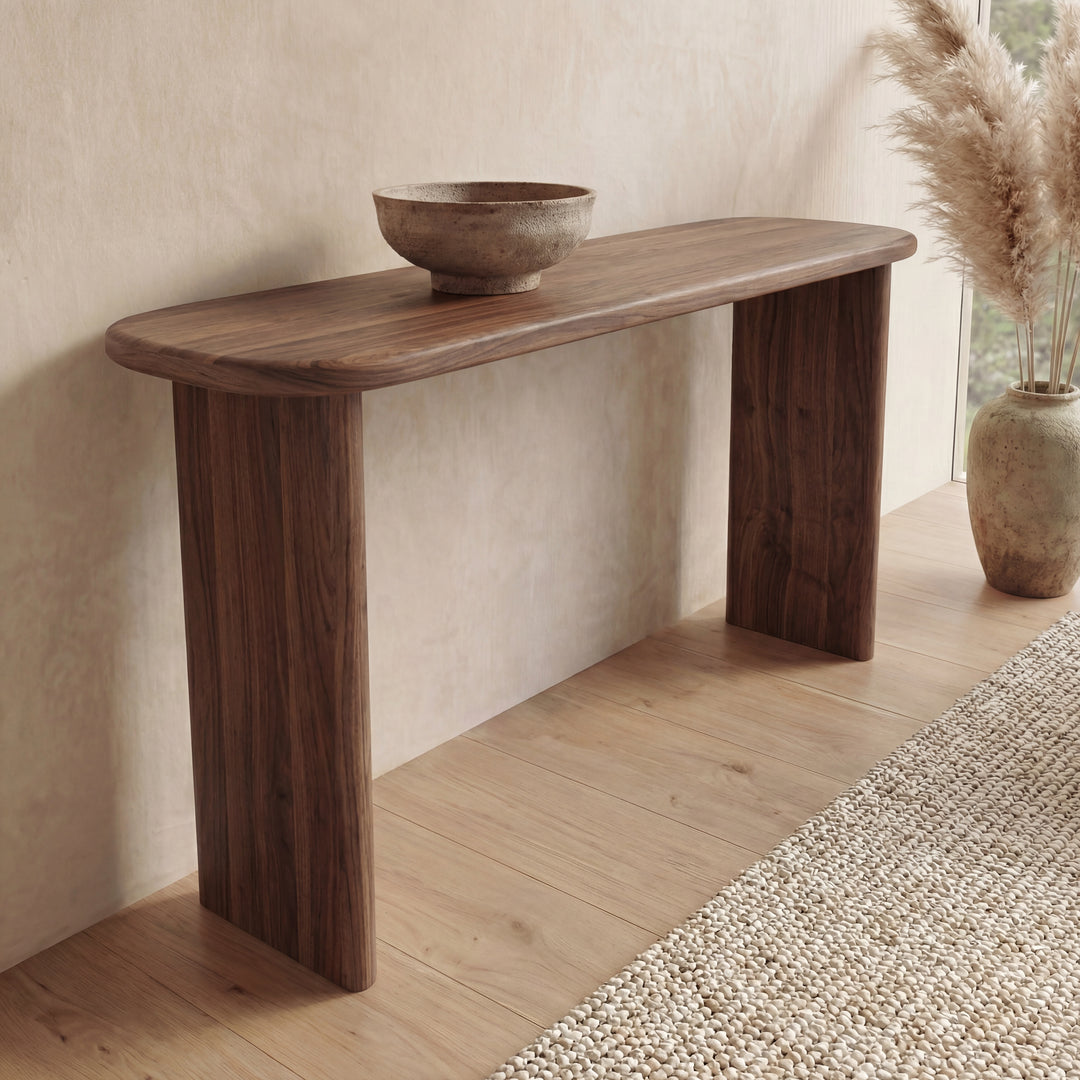 The 60 inch console table in walnut, handmade in Vancouver with rounded edges, displays a rustic wooden bowl. It stands on a light wood floor by a textured rug, while a large vase with pampas grass is placed near the window.