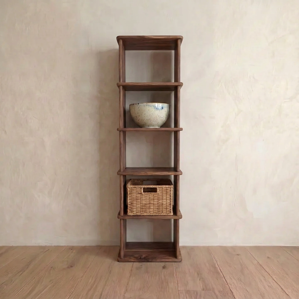 A narrow bookshelf in walnut with four shelves stands against a beige textured wall. A ceramic bowl is placed on the second shelf and a wicker basket sits on the third, while the floor beneath is wooden.