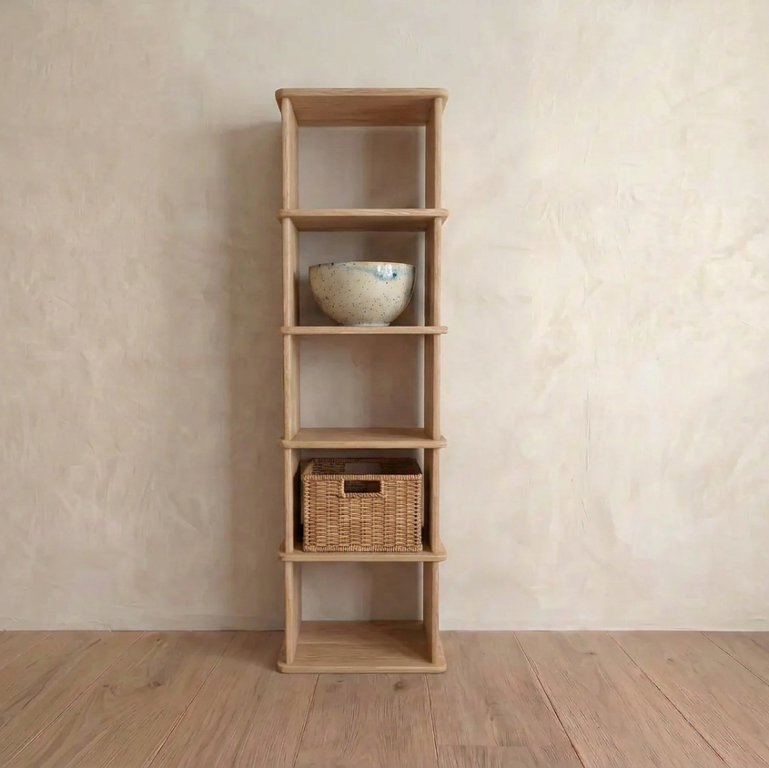 A narrow bookshelf in white oak stands against a light beige wall, with a ceramic bowl on the second shelf from the top and a woven wicker basket on the fourth. The remaining shelves are empty.