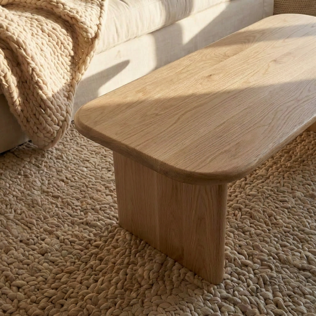 A 40 inch coffee table in white oak sits on a chunky woven beige rug beside a sofa topped with a thick knitted blanket, all illuminated by warm natural sunlight.