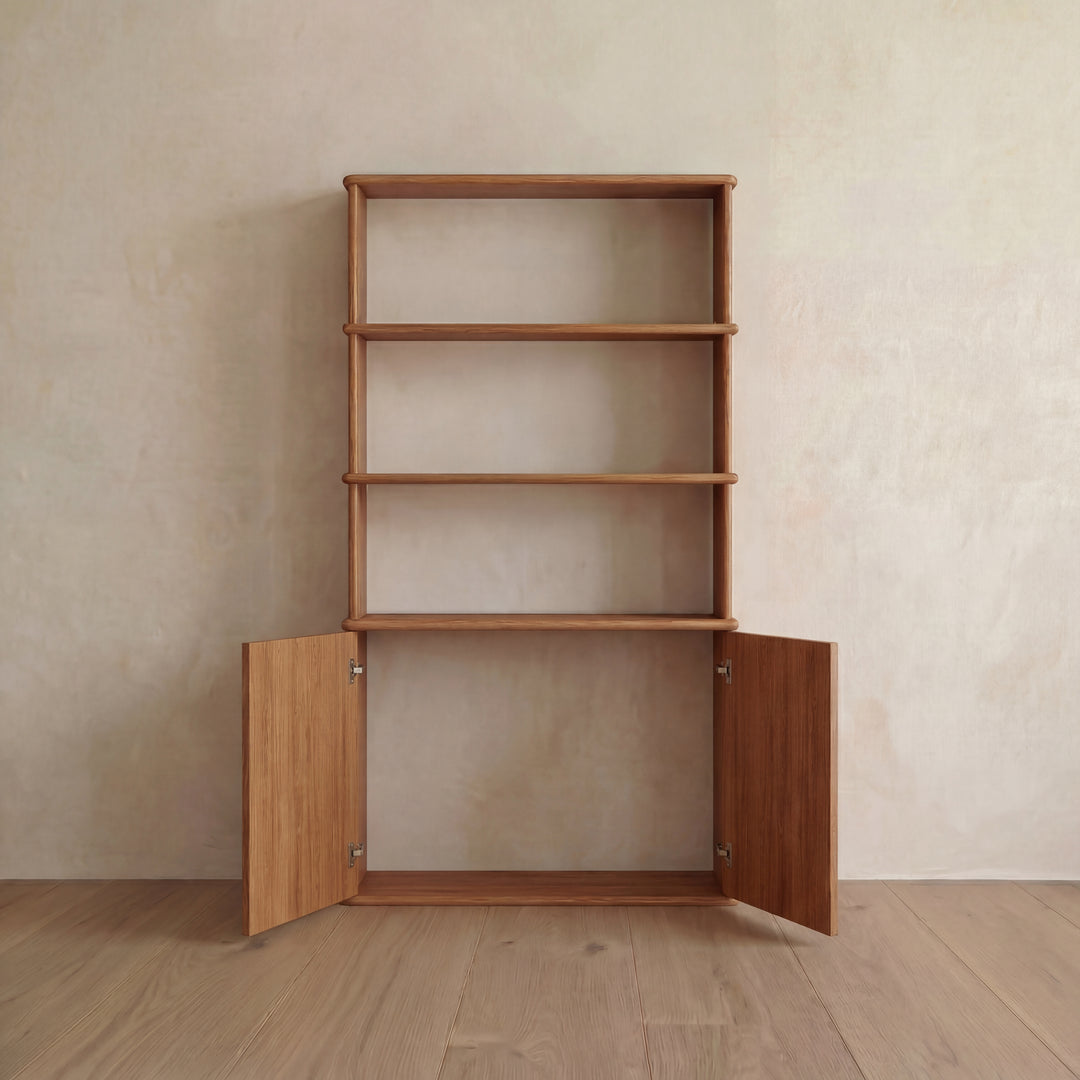 The Cabinet bookshelf in red oak stands against a beige wall on a wooden floor in this Vancouver-inspired setting. This shelving unit features three open shelves above and a two-door cabinet below, thoughtfully designed with push-to-open doors for seamless access.