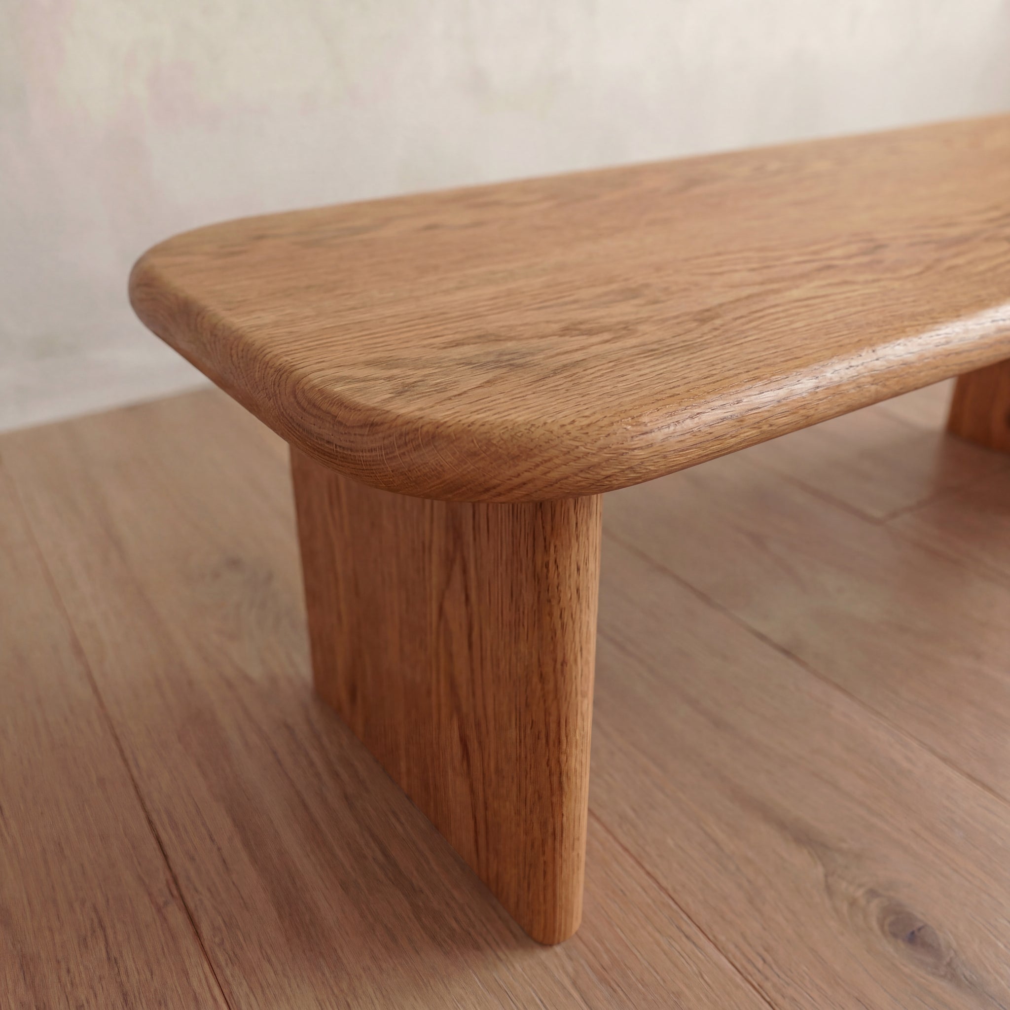 A close-up view of a 60 inch coffee table in red oak, featuring rounded edges and sturdy legs, crafted from solid wood, placed on a light wood floor against a neutral wall background.