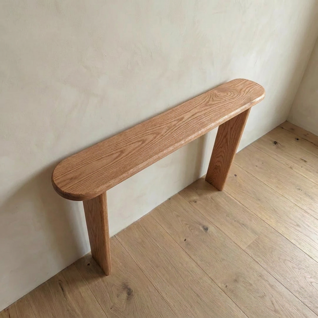 A minimalist narrow console table in red oak, handcrafted in Vancouver with two legs, features a simple elongated oval top and sits against a light wall on a wooden floor.