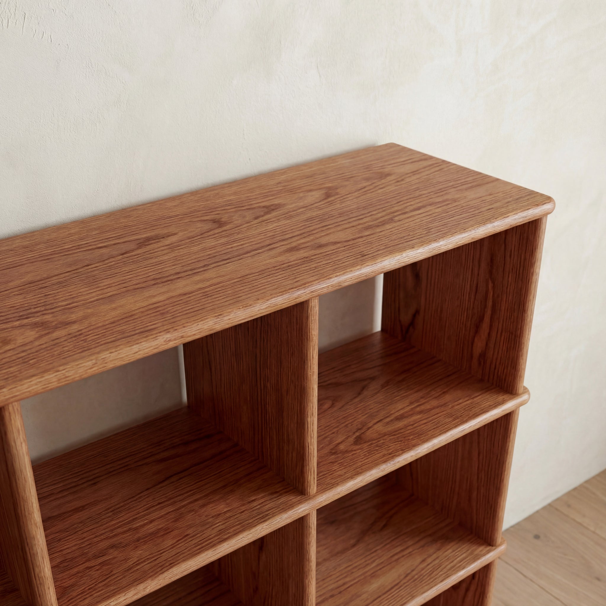 The Cube bookshelf in red oak features a handmade natural finish, three shelves, and multiple square compartments. It's shown against a light-colored textured wall.