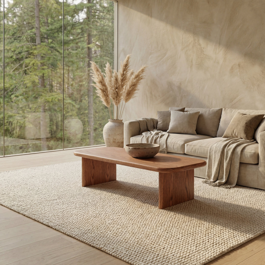 A modern living room featuring a beige sofa, neutral cushions, and a handmade 60 inch coffee table in red oak. A textured rug and pampas grass in a large vase complete the space, all framed by floor-to-ceiling windows overlooking a serene forest.