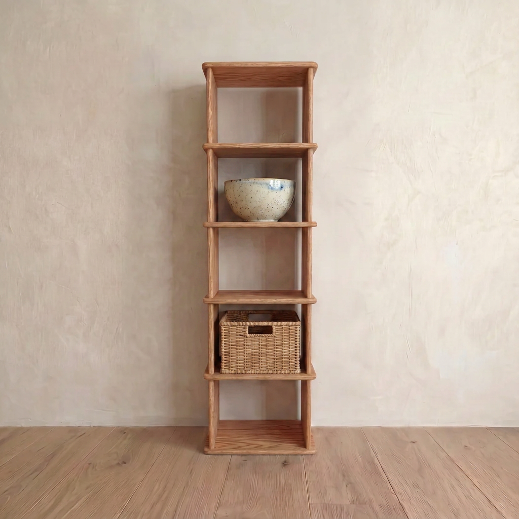 A narrow bookshelf in red oak stands against a light wall. On its middle shelf rests a handmade speckled ceramic bowl, and below, a square wicker basket sits on the wooden floor.