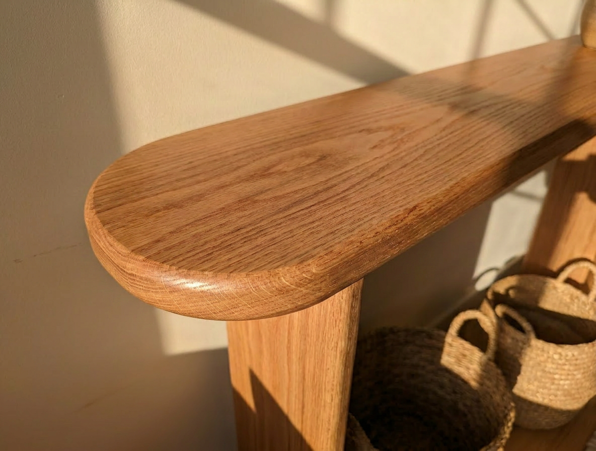 A close-up of the Narrow console table in red oak with a natural finish. Sunlight casts shadows on its smooth, rounded surface. Two woven baskets rest on a lower shelf. This handmade piece is crafted in Vancouver.
