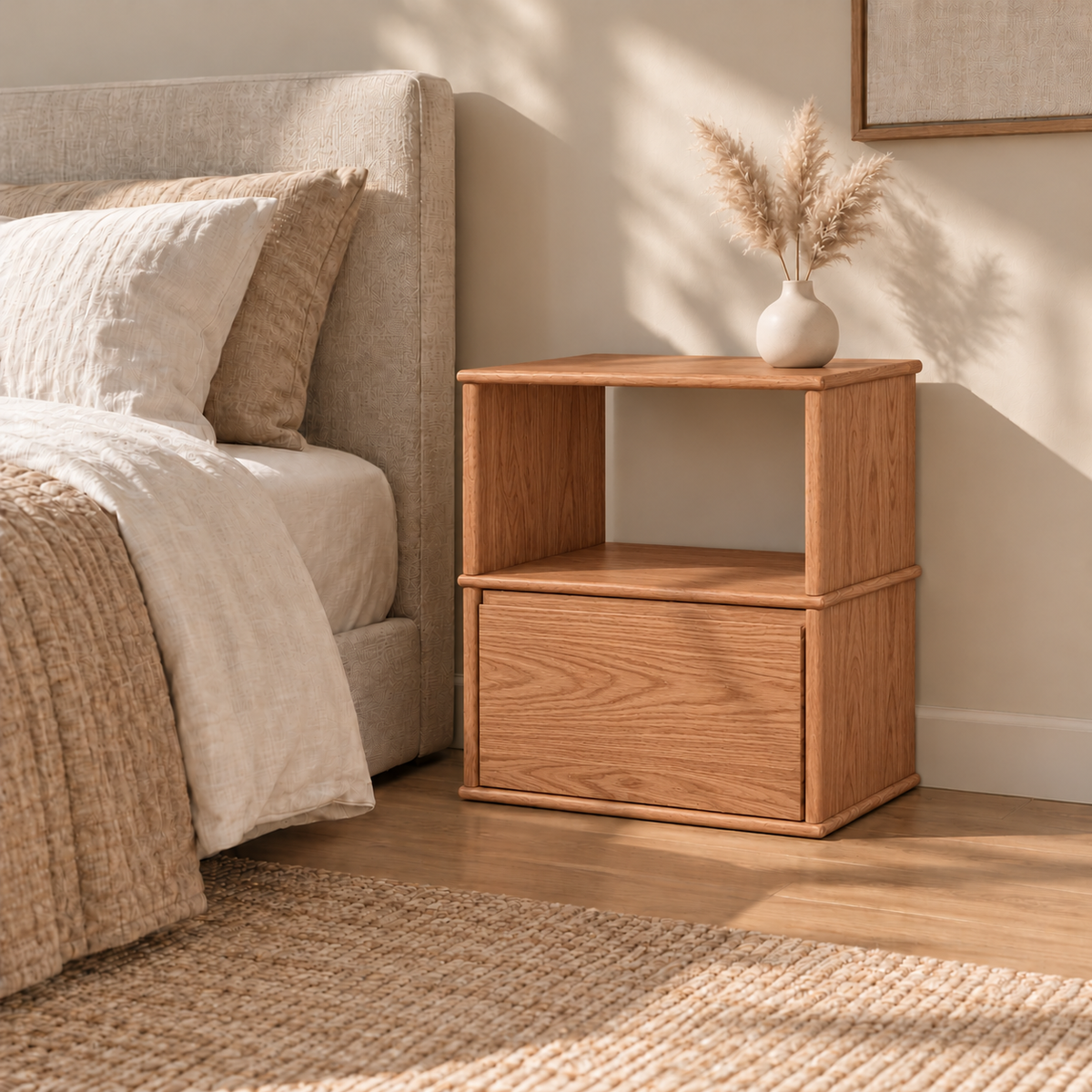 A solid wood nightstand in red oak beside a bed with linen bedding, with a vase of pampas grass on top and a jute rug below.