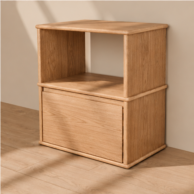 A solid wood nightstand in white oak with an open shelf above a single drawer and softly rounded edges, against a plain plaster wall.