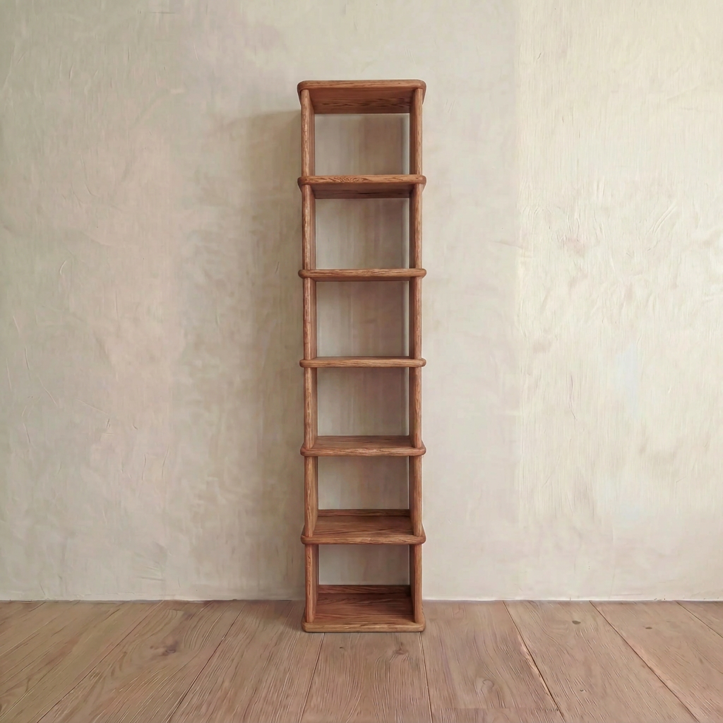 The Tall Narrow Bookcase in Red Oak features six shelves and a minimalist design. Handmade from red oak, it stands empty against a light textured wall on a wooden floor, offering an elegant and understated storage solution.