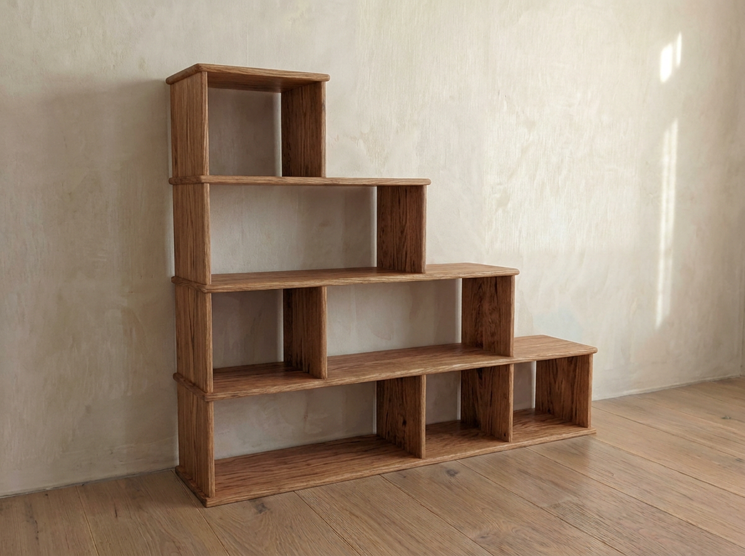 The Step bookshelf in red oak features four handmade, tiered shelves rising from right to left like stairs, and stands elegantly against a light wall atop a wooden floor.