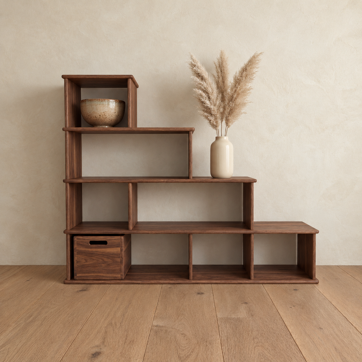The step bookshelf in walnut against a plaster wall, with a bowl in the top cube, a vase of pampas grass on a step and a wooden storage cube in the bottom row.