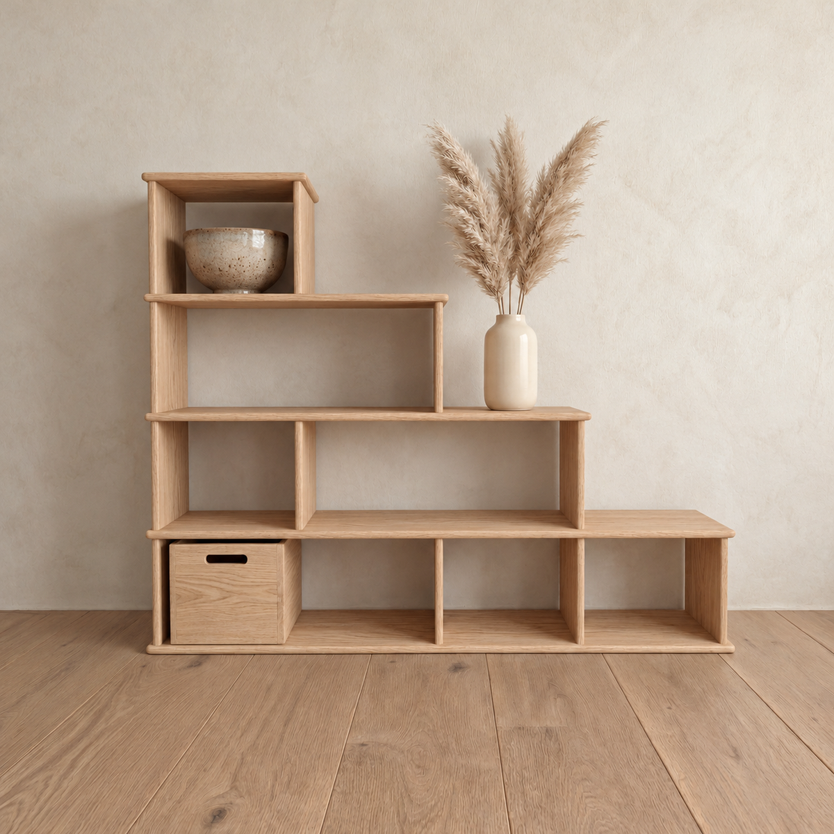 The step bookshelf in white oak against a plaster wall, with a bowl in the top cube, a vase of pampas grass on a step and a wooden storage cube in the bottom row.