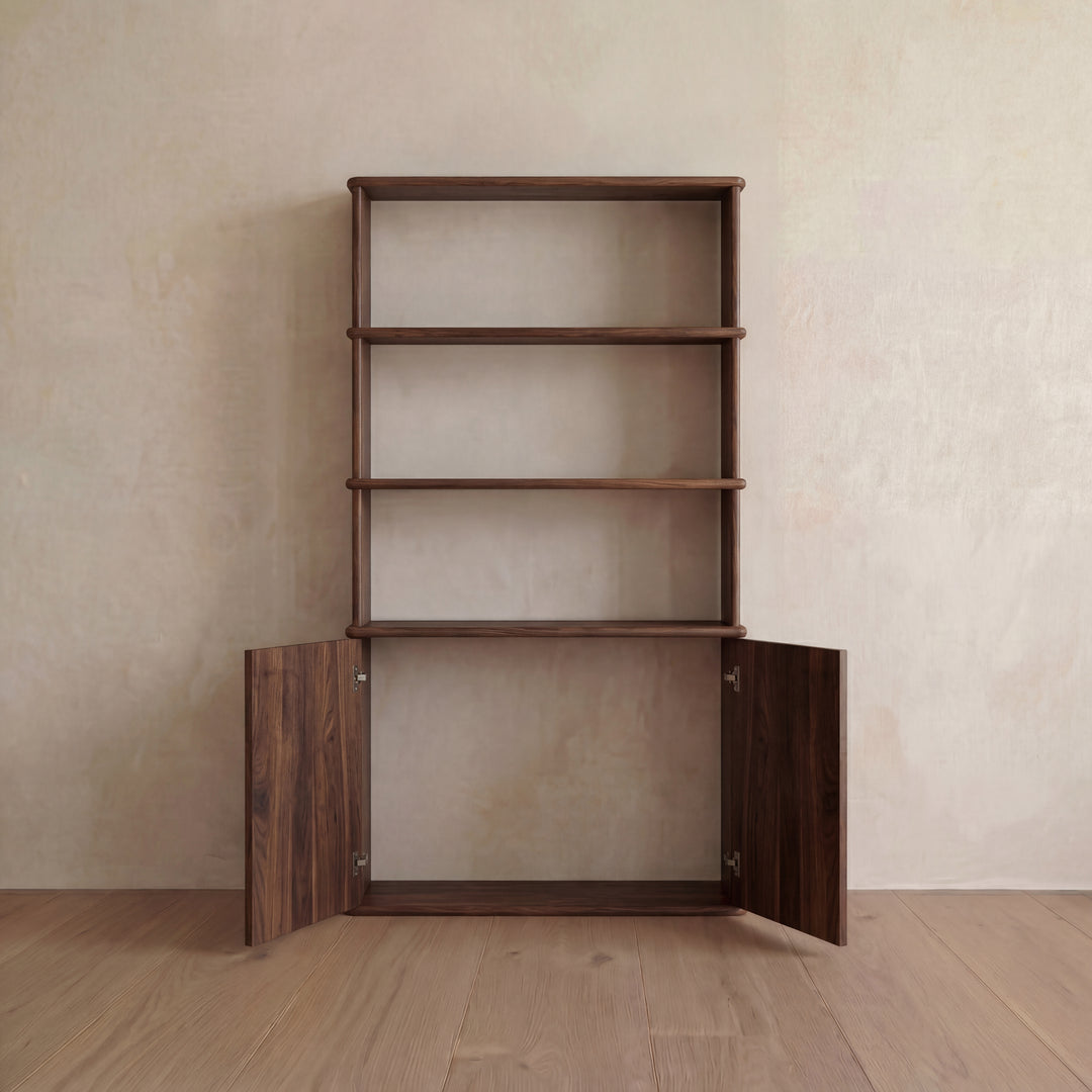 The Cabinet bookshelf in walnut, which features three open shelves and a bottom cabinet with push-to-open doors, stands against a light beige wall on a wooden floor.
