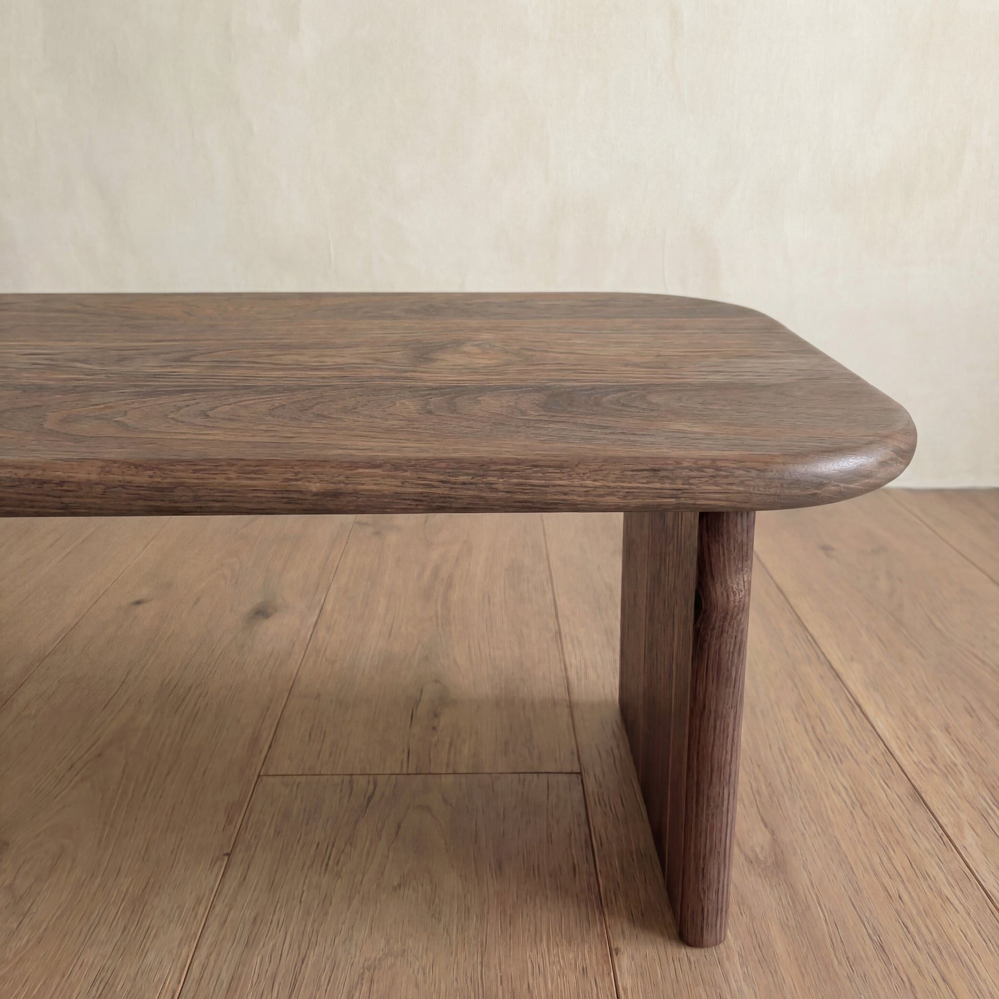 A close-up of the 60 inch coffee table in walnut, featuring rounded edges and a single sturdy leg, set on a wood floor against a beige wall.