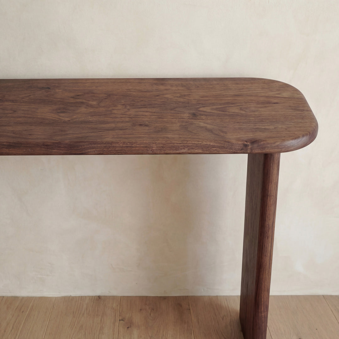 The 60 inch console table in walnut, handmade in Vancouver, displays a wooden tabletop with rounded corners and a smooth finish. Seen against a light beige wall on a wood floor, only the tabletop and part of one leg are visible.