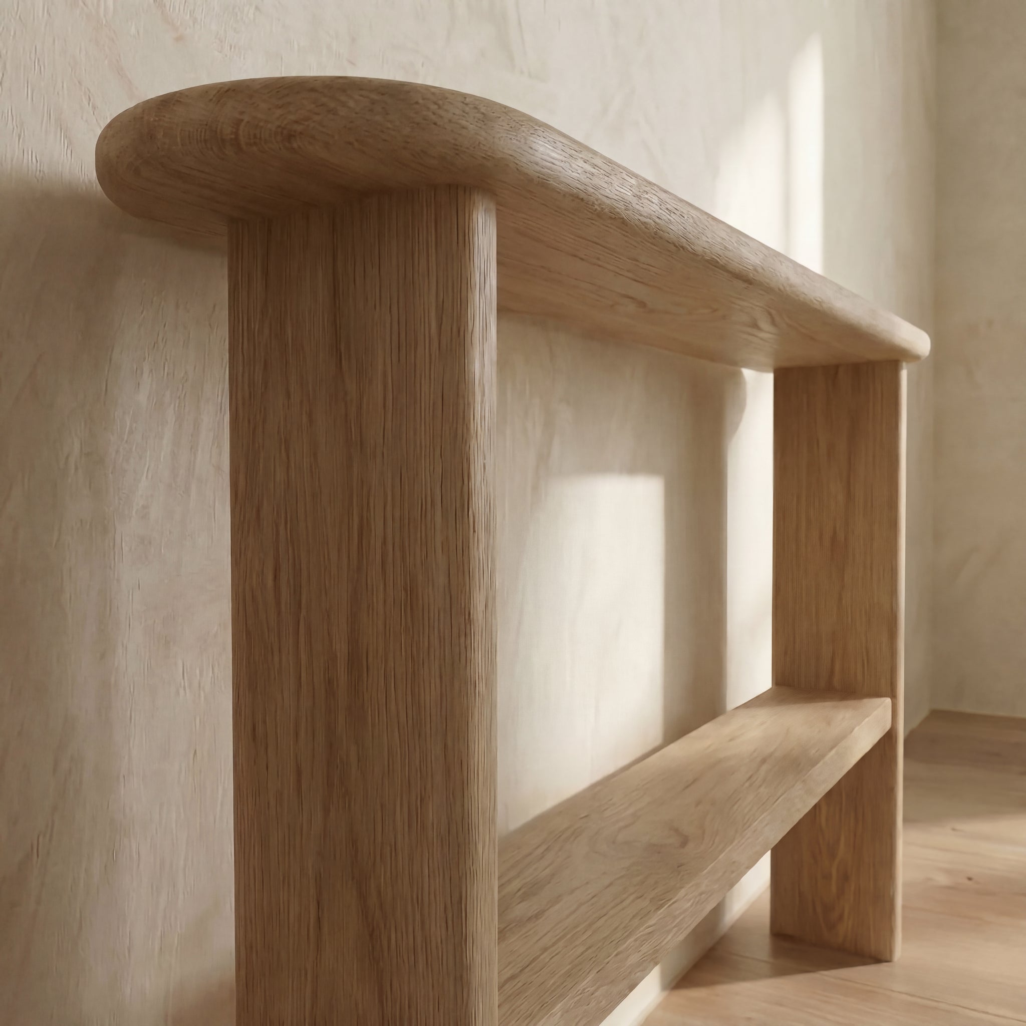 A white oak console table with a lower shelf stands against a light-colored wall, its minimalist design and rounded edges highlighted by natural sunlight.
