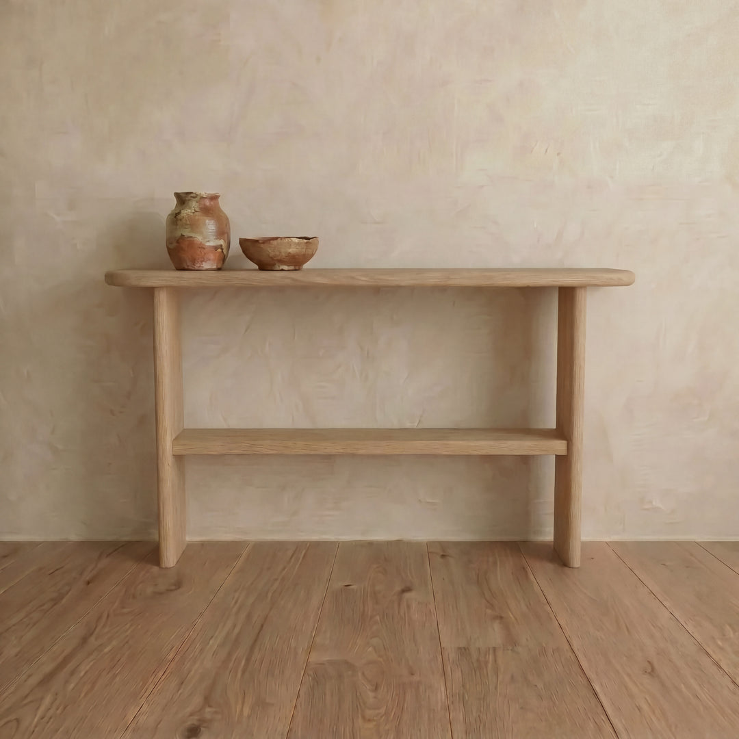 A Console table with shelf in white oak stands against a textured beige wall. Two rustic ceramic vessels sit on top, enhancing the minimalist look and blending with the natural wood flooring.