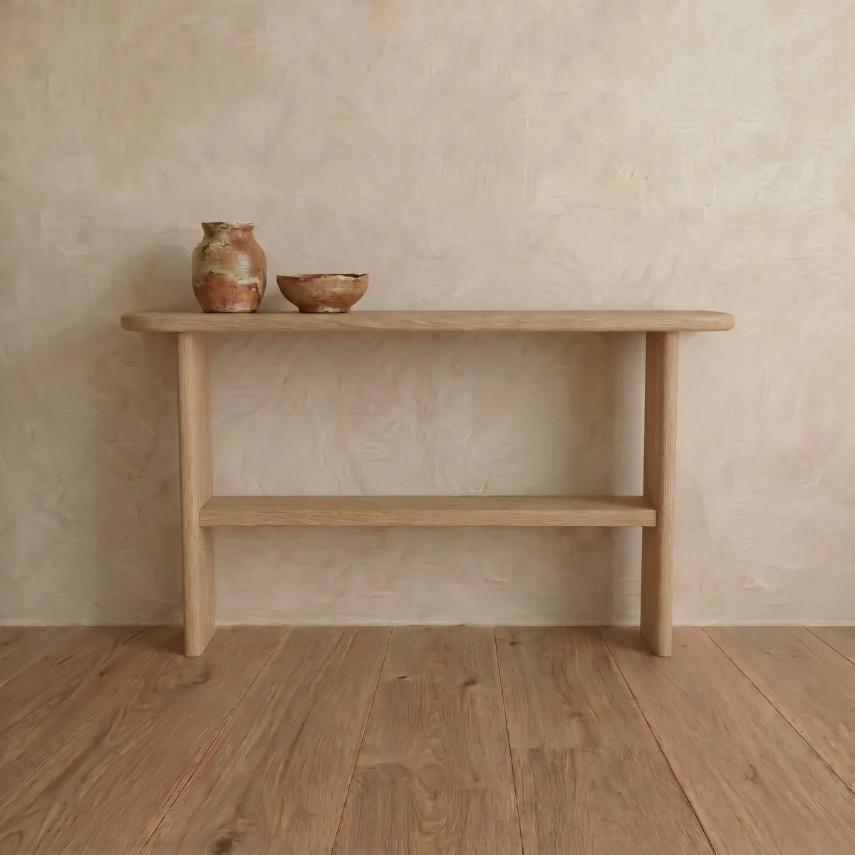 A Console table with shelf in white oak stands against a textured beige wall. Two rustic ceramic vessels sit on top, enhancing the minimalist look and blending with the natural wood flooring.